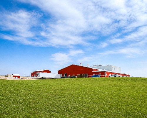 Blue sky, white clouds, green grass, red side of building, white roof, trailer, Upper Iowa Beef plant in 2018.