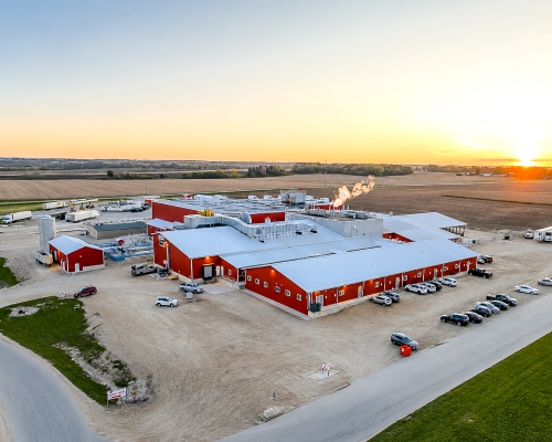 Sunrise over Upper Iowa Beef beef processing plant, blue and yellow skies, green grass, cars in parking lot, Casey's, red siding and white roof.