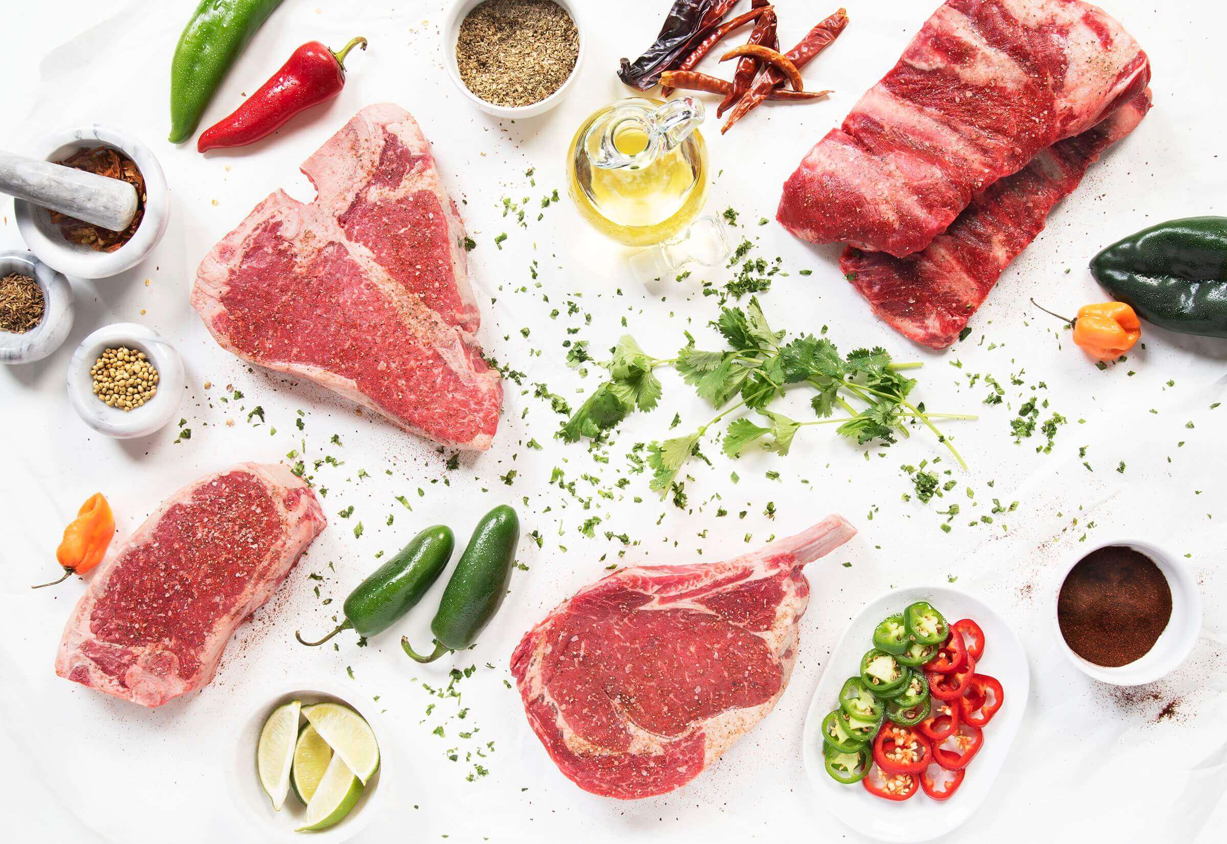 Assorted raw steaks with chili peppers, spices, herbs, lime wedges, and oil on a white background.