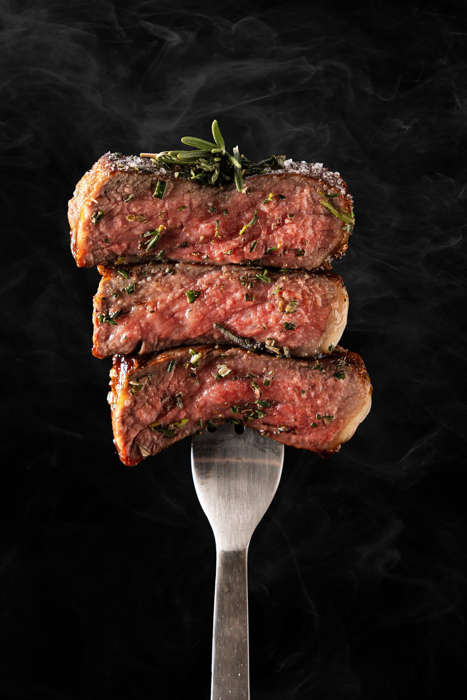 Three medium-rare steak slices with herbs stacked on a fork against a smoky black background.