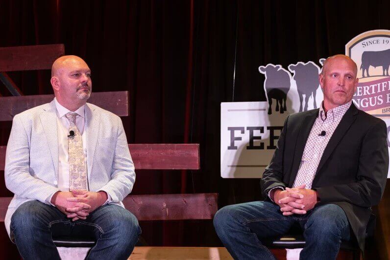 Two men in suits sit on stage with cattle-themed signs in the background, facing the audience.