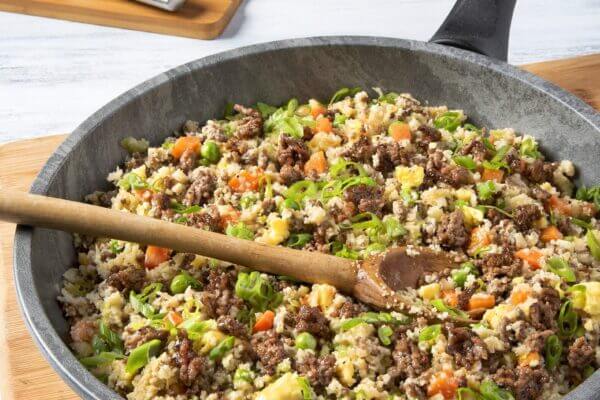A skillet of ground beef fried rice with vegetables and a wooden spoon, on a wooden cutting board.