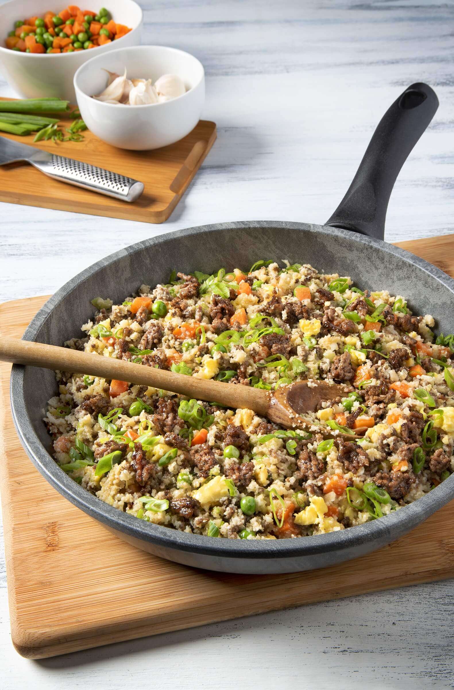 A skillet of ground beef fried rice with vegetables and a wooden spoon, on a wooden cutting board.
