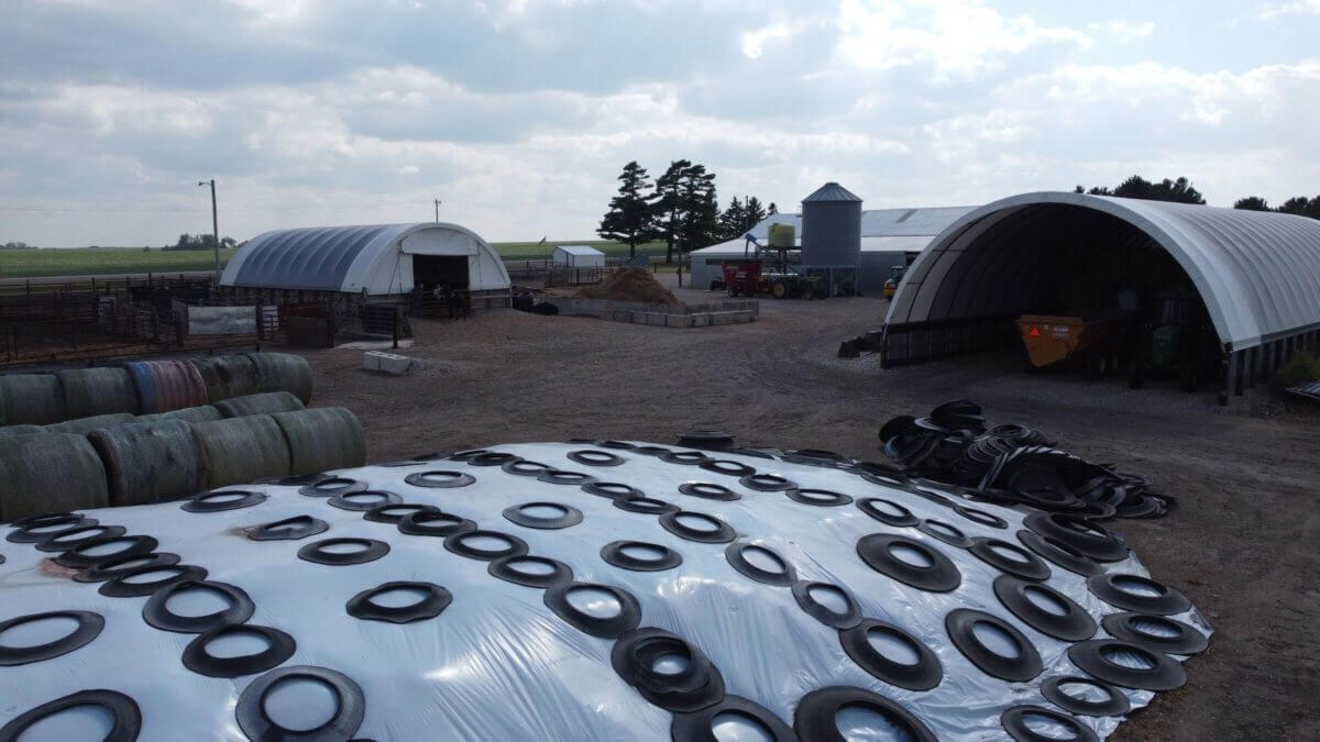 A farmyard with silage covered by plastic and tires, barns, hay bales, machinery, and a silo in view.