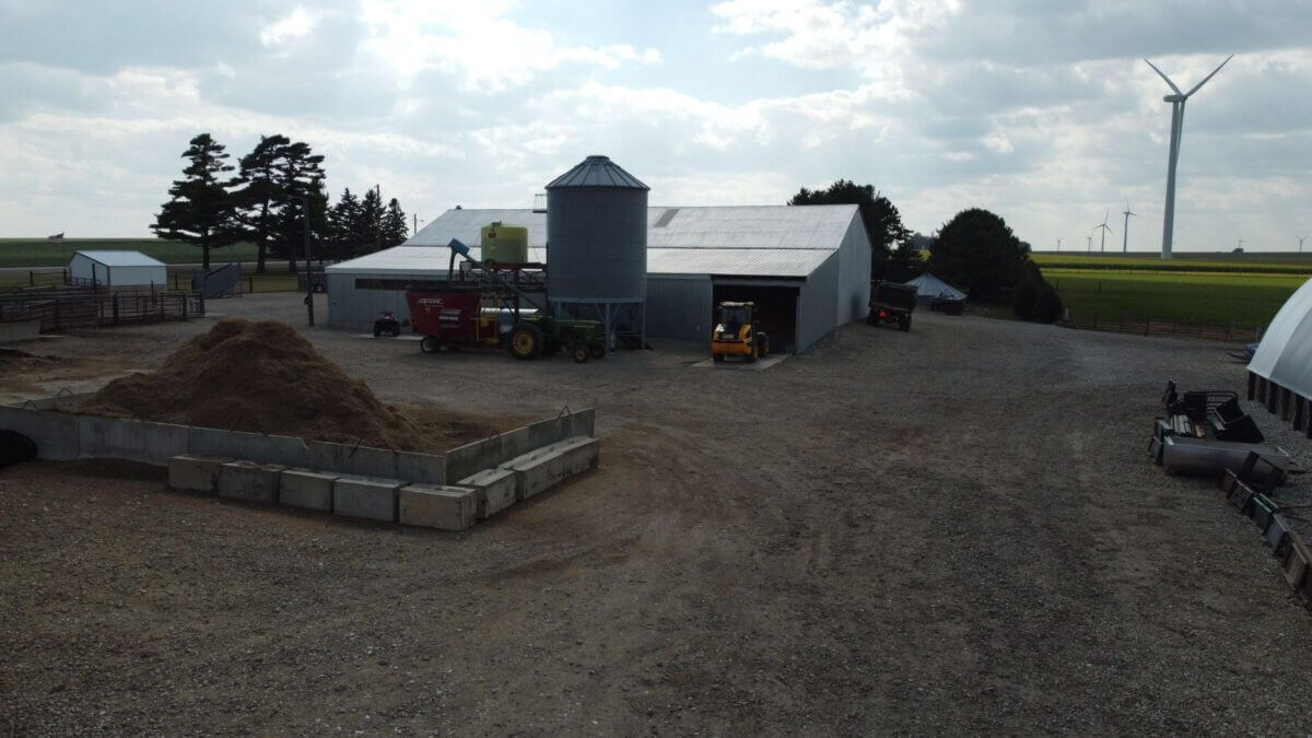 A farmyard with barns, a silo, tractors, a feed pile, and a wind turbine in the background.