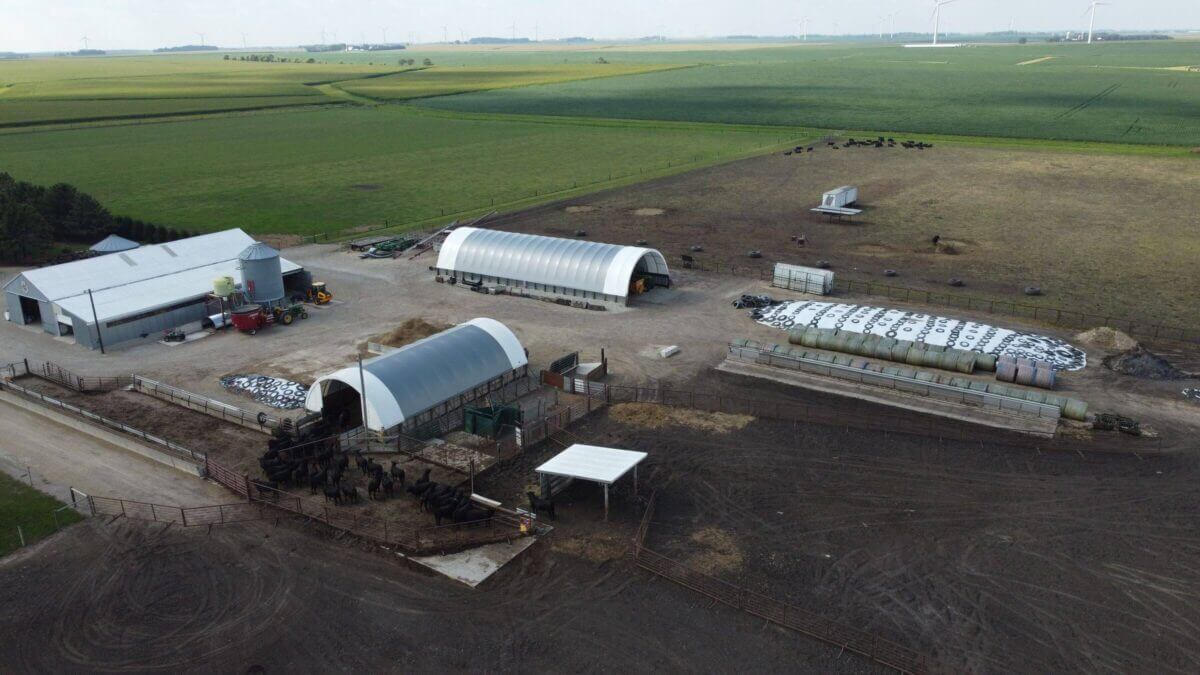 Aerial view of a farm with barns, livestock, machinery, hay bales, and fenced enclosures on green fields.