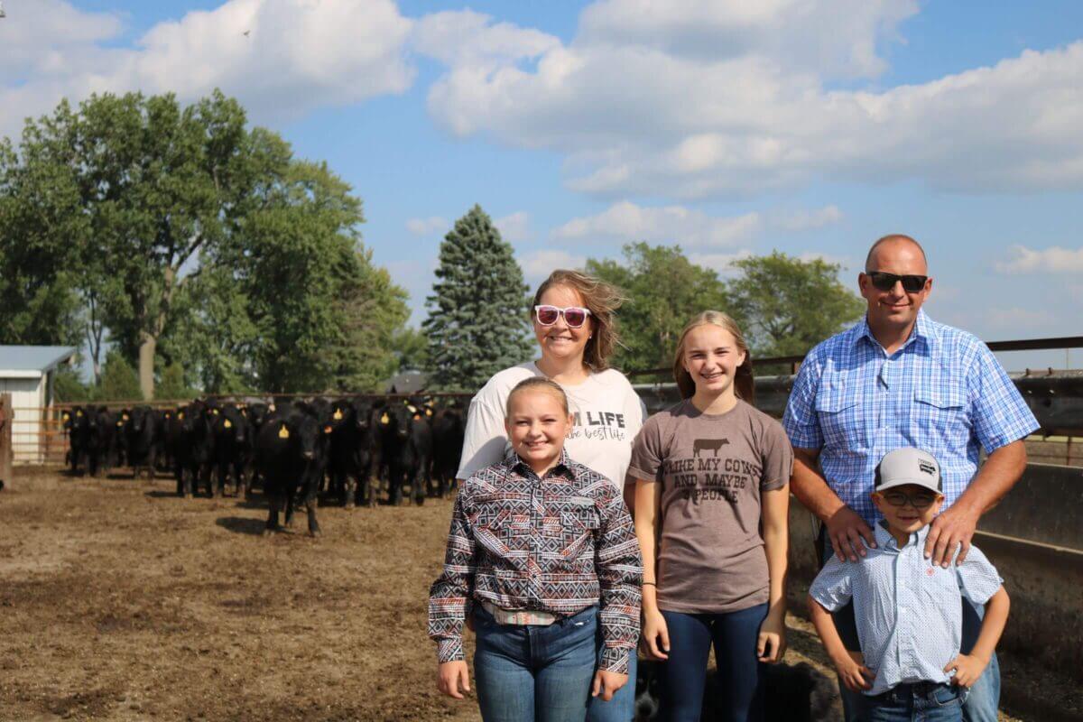A family of five poses in front of a herd of black cows on a sunny day at a farm.