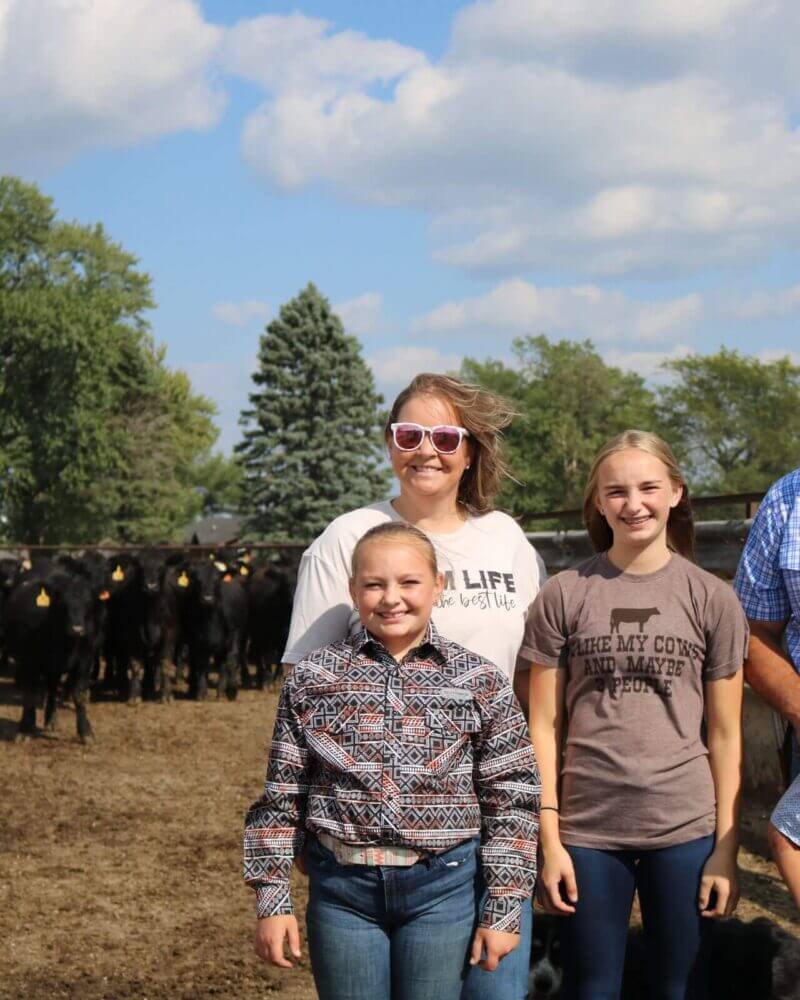 A family of five poses in front of a herd of black cows on a sunny day at a farm.