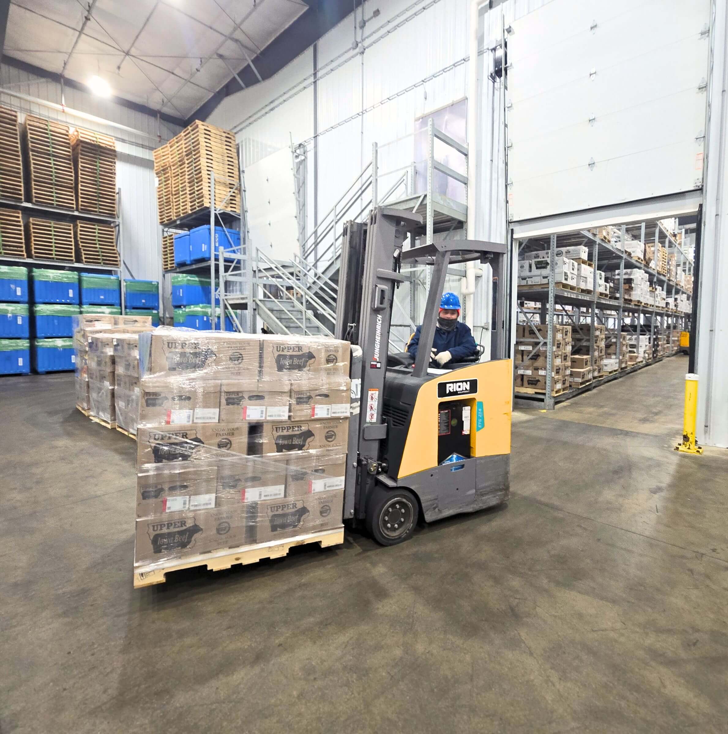 A worker drives a forklift moving boxes on a pallet in a warehouse with shelves and stacked pallets.