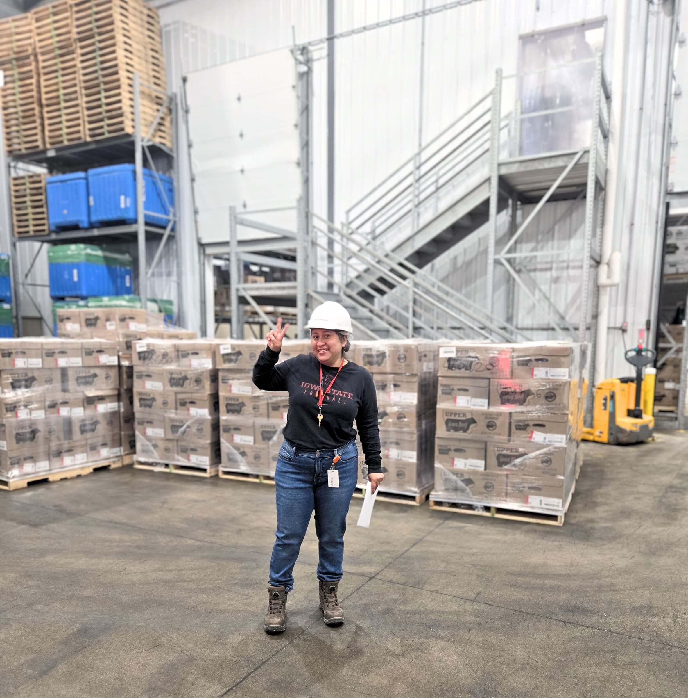 A person in safety gear stands in a warehouse, smiling and making a peace sign.
