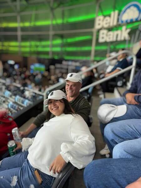 A smiling couple in white hats sit in stadium seats with a "Bell Bank" sign in the background.