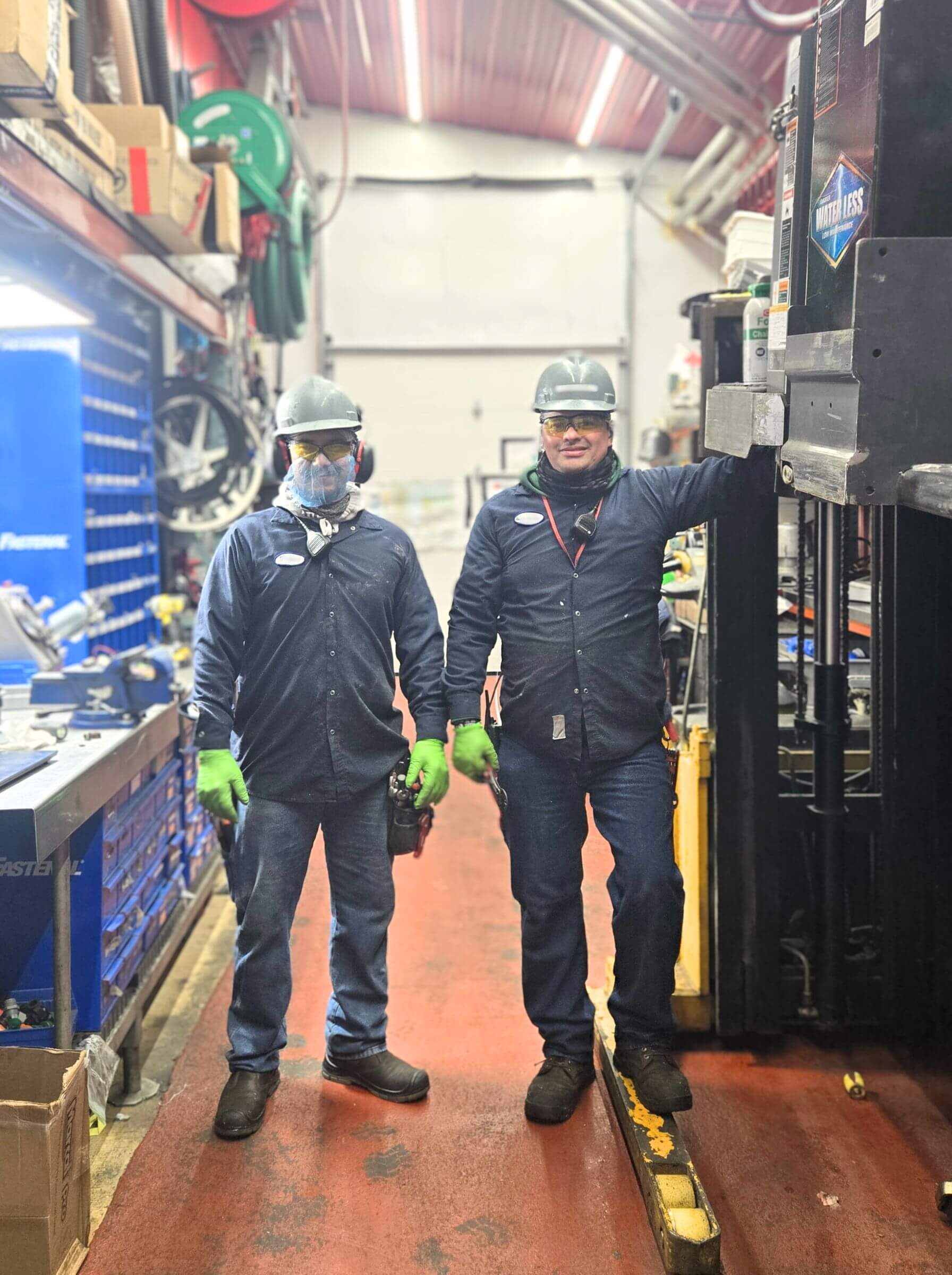 Two workers in safety gear stand in a workshop with tools and equipment visible around them.