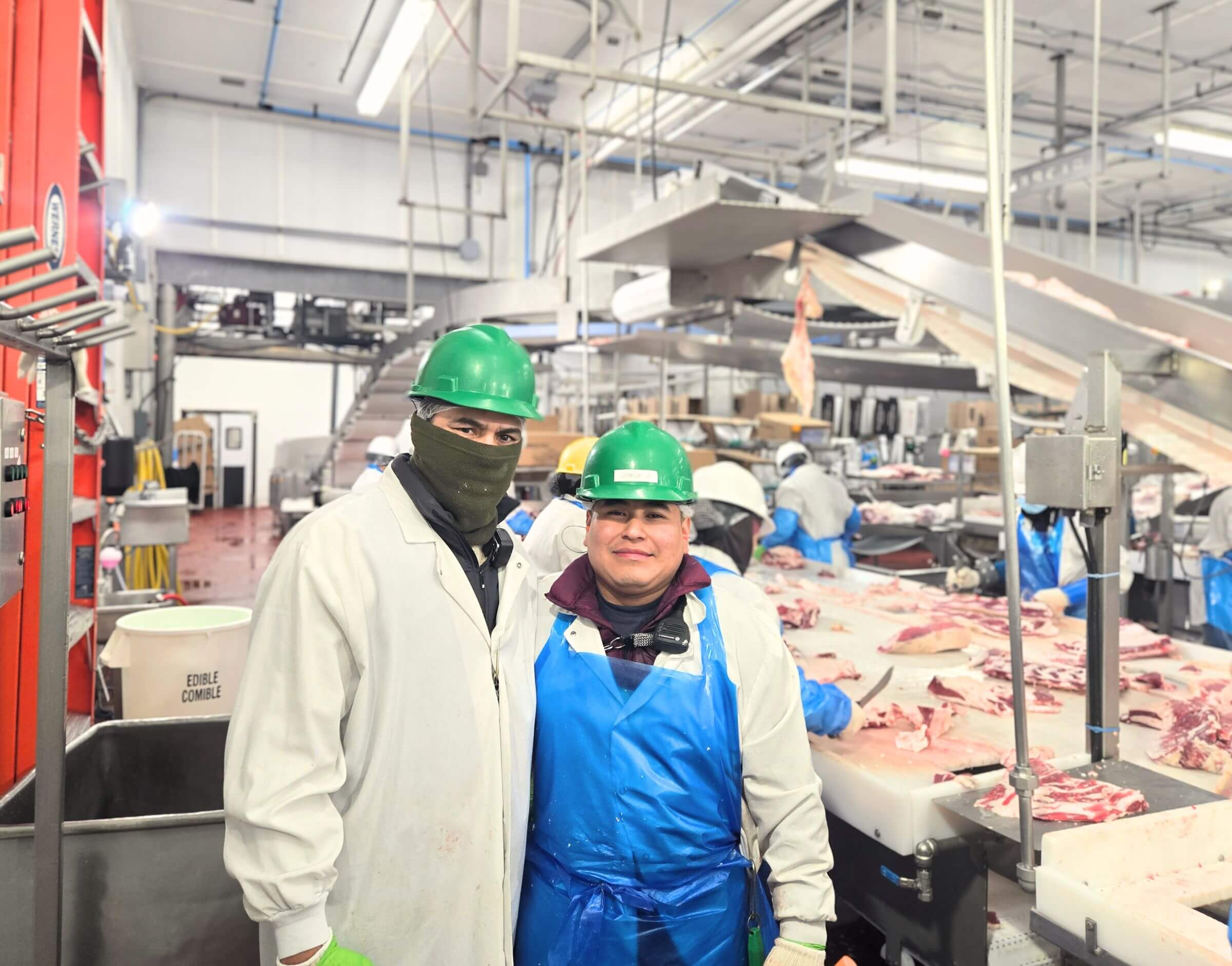 Two workers in protective gear and green helmets pose in a meat processing facility with meat on tables.