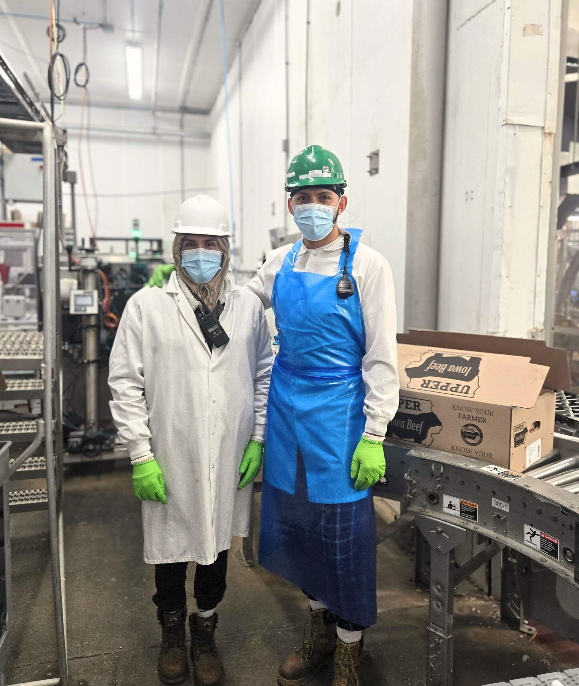 Two workers in protective gear, masks, and gloves stand in a food processing facility next to a cardboard box.