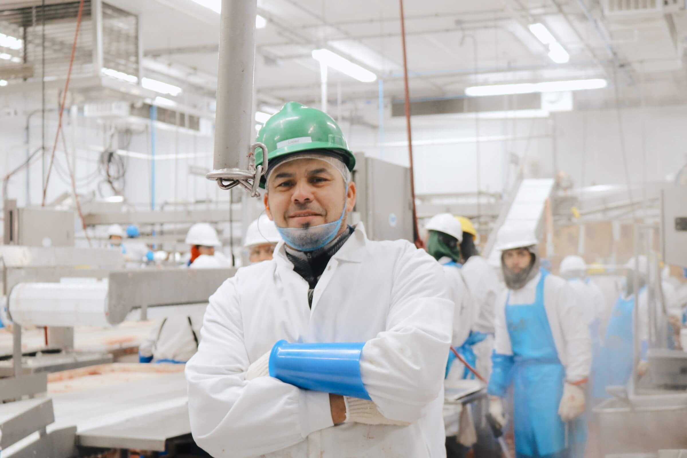 Worker in a green hard hat and white coat stands with arms crossed in a busy food processing facility.