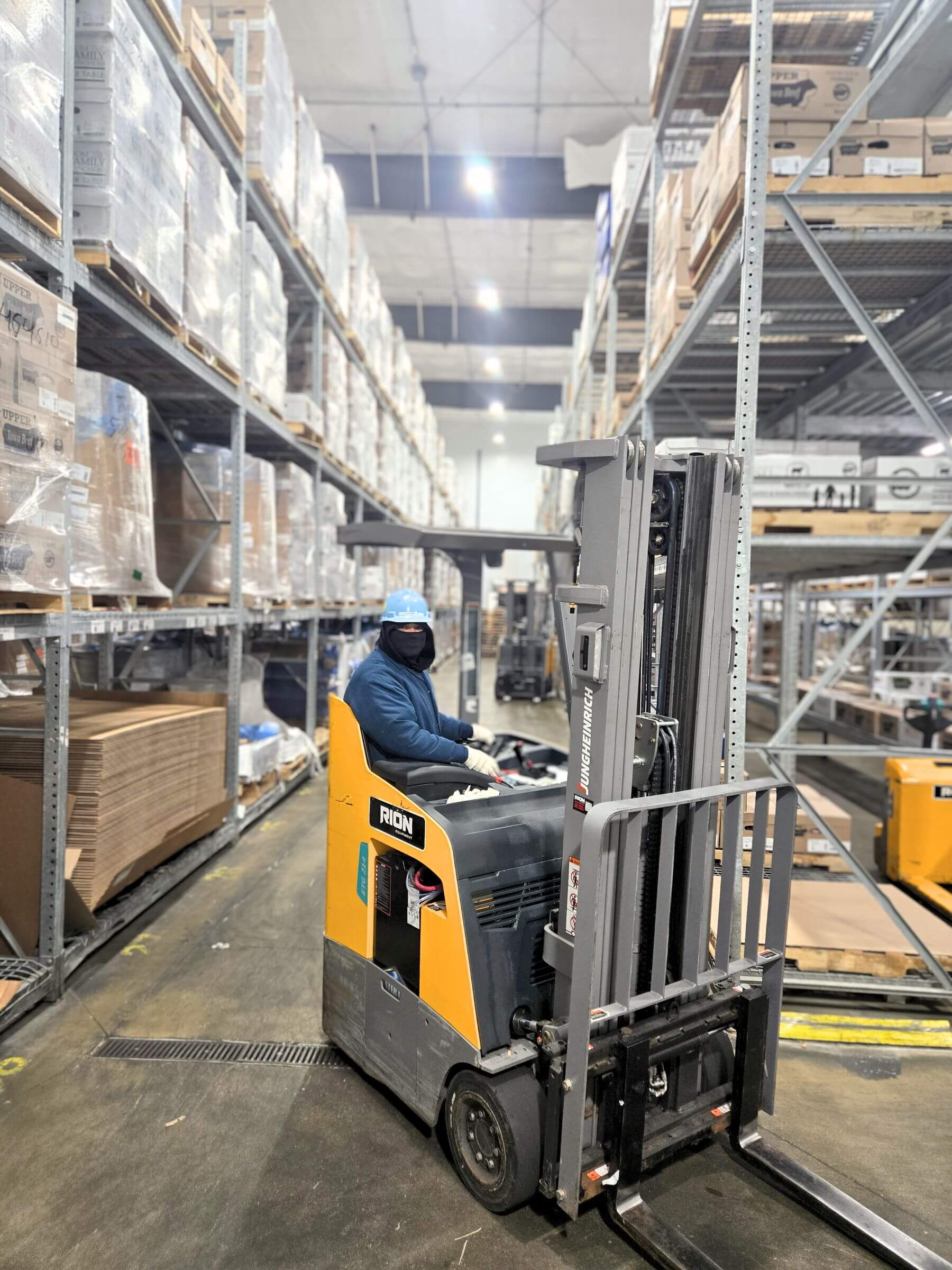 A person wearing a mask operates a forklift in a large warehouse filled with stacked pallets.