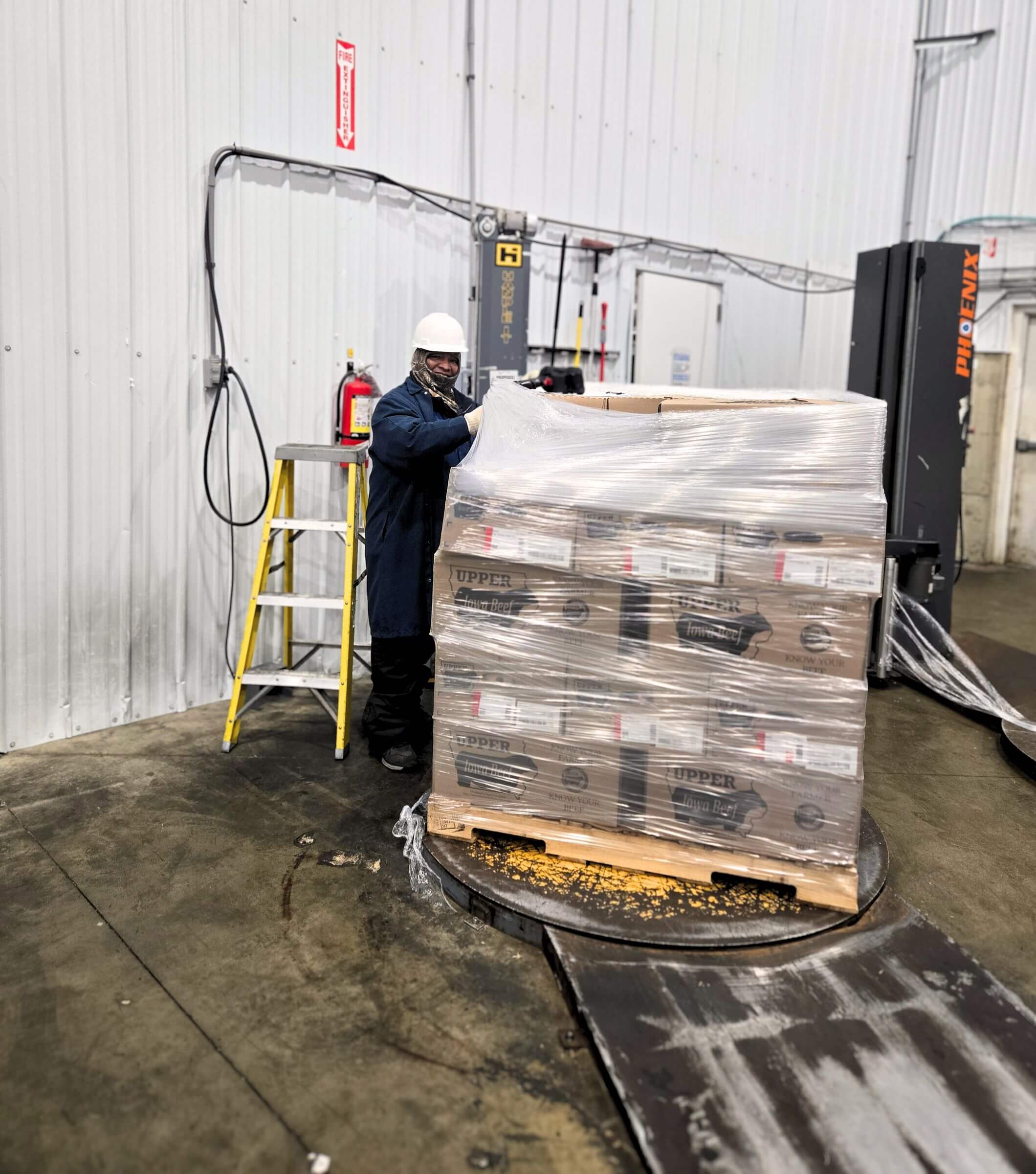 Worker wrapping a pallet of boxed goods in plastic near a ladder and fire extinguisher in a warehouse.
