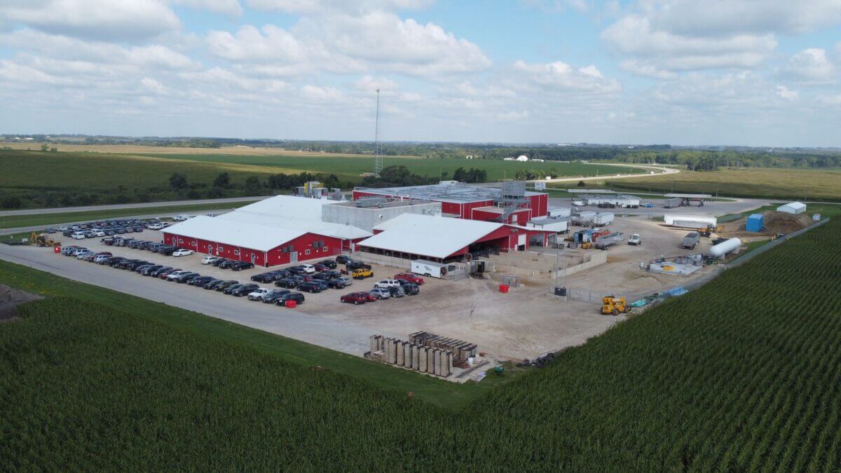 Aerial view of a large red industrial building surrounded by parked cars and farmland under a partly cloudy sky.
