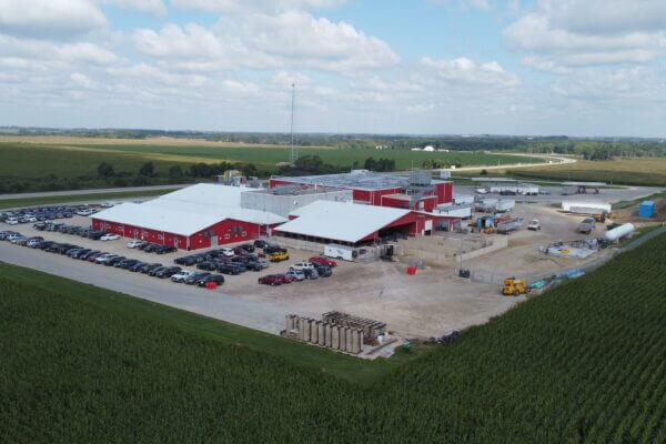 Aerial view of a large red industrial building surrounded by parked cars and farmland under a partly cloudy sky.