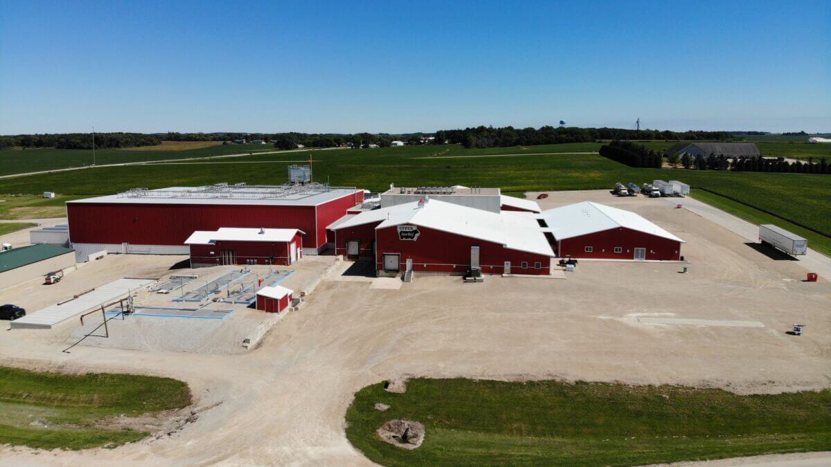 Aerial view of a large red farm building complex surrounded by green fields under a clear blue sky.
