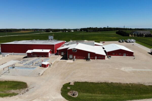 Aerial view of a large red farm building complex surrounded by green fields under a clear blue sky.