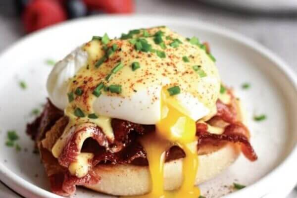 A poached egg on bacon and an English muffin with chives on top, yolk running, on a white plate.