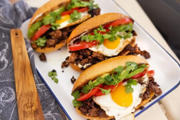 Beef Chorizo Sausage, green fresh cilantro, sunny-side up egg, white buns, sliced red tomato, sliced green avocado, white serving plate.