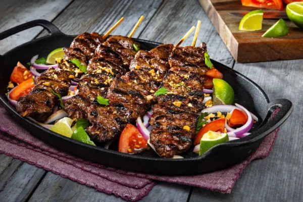 Beef Suya (Grilled Beef Skewers), red tomatoes, red onions sliced, limes, corn, cast iron, picnic table, seasonings.