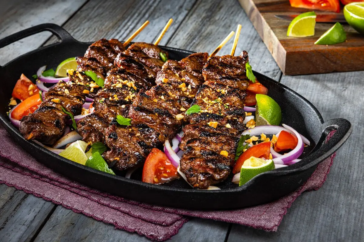 Beef Suya (Grilled Beef Skewers), red tomatoes, red onions sliced, limes, corn, cast iron, picnic table, seasonings.