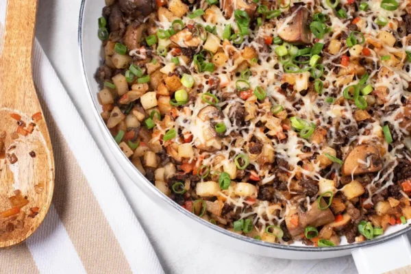 Beef Swiss and Mushroom Hash, wooden spoon laid on white and tan striped towel next to white pan ith ground beef, cheese, hashbrown, diced veggies, and sliced green onions.