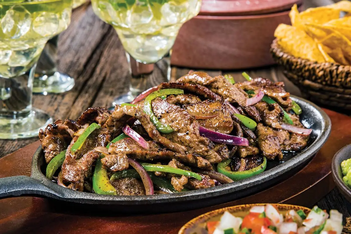 Flank Steak Fajitas, sliced avocadoes, sliced red onion, cast iron skillet on wooden table with margaritas.