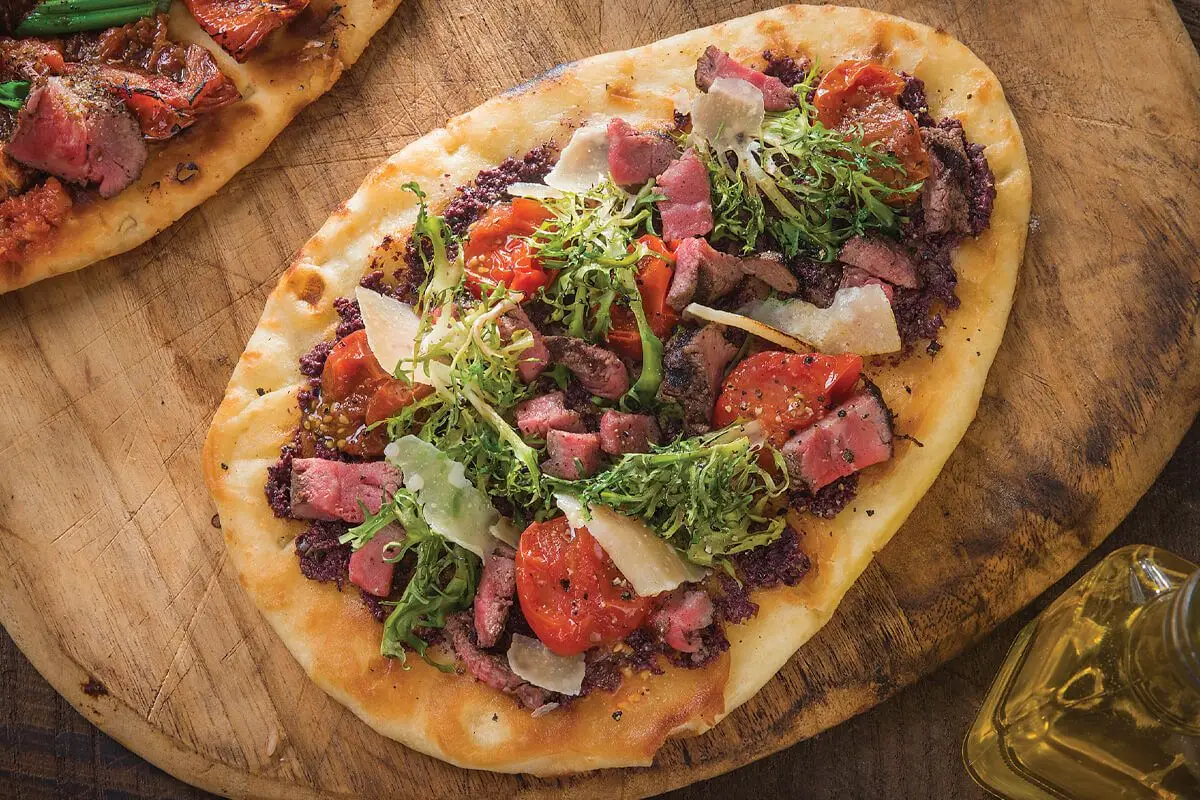 Mediterranean Steak Flatbread, red sliced tomatoes, green lettuce, cheese and steak slices, wooden cutting board.