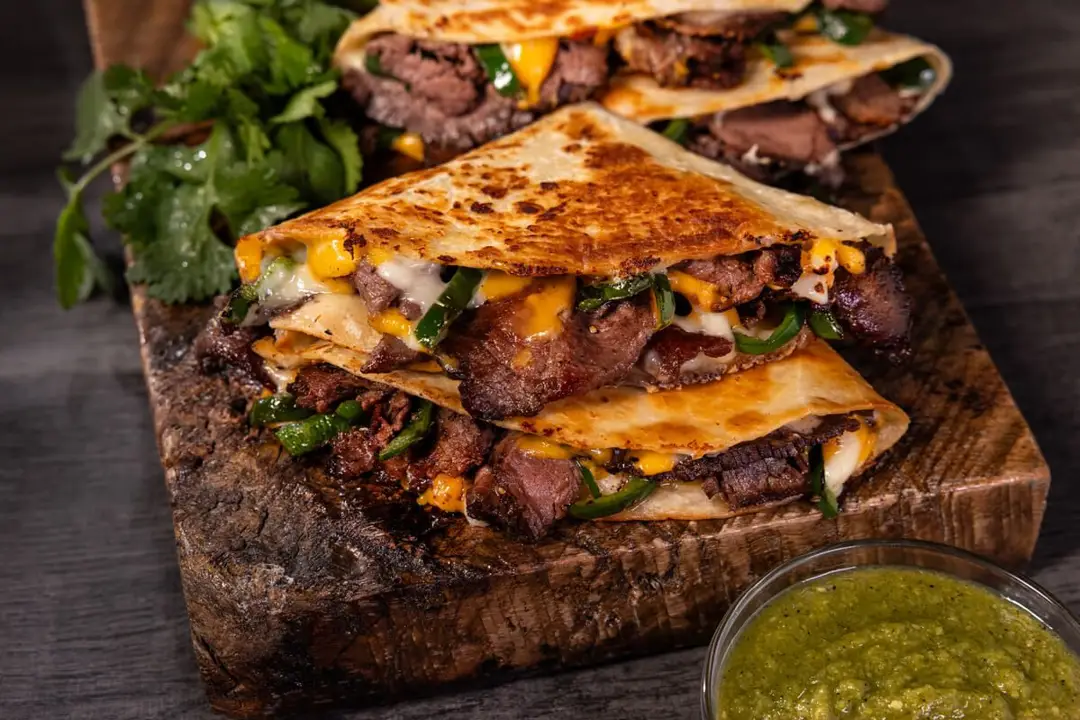 Roast Beef Fajita Quesadilla, fried soft taco shell, greens and cheese on wooden tray.